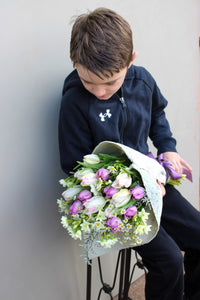 A young boy in a black tracksuit sits holding Wild About Flowers' Spring Cheer Bunch, a mix of purple and white tulips wrapped in decorative paper. He leans slightly, gazing down at the seasonal tulips against a neutral background.