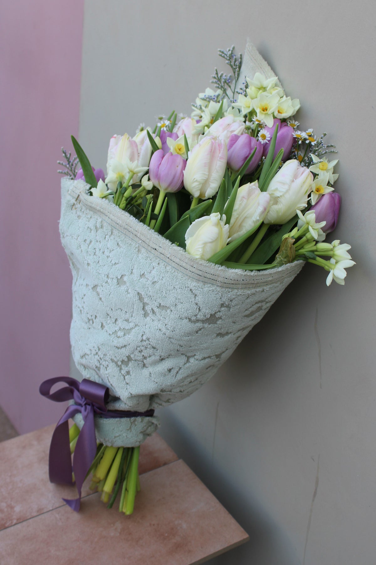 The Spring Cheer Bunch from Wild About Flowers features white and purple tulips mixed with small white blooms, wrapped in sustainable fabric, tied with a purple ribbon, and leaning elegantly against a tiled wall.