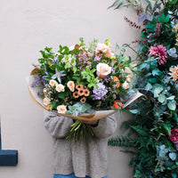 A person in a gray sweater holds 'The Sunday' hand-tied spring bouquet by Wild About Flowers, with peach, purple, and pink blooms, standing near a flower- and greenery-decorated wall.