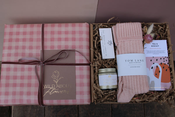 The 'Valentines Night in' Gift Box by Wild About Flowers is open, showing alpaca wool bed socks, tea, a candle, chocolate, dried flowers, and a skincare item arranged on shredded brown paper.