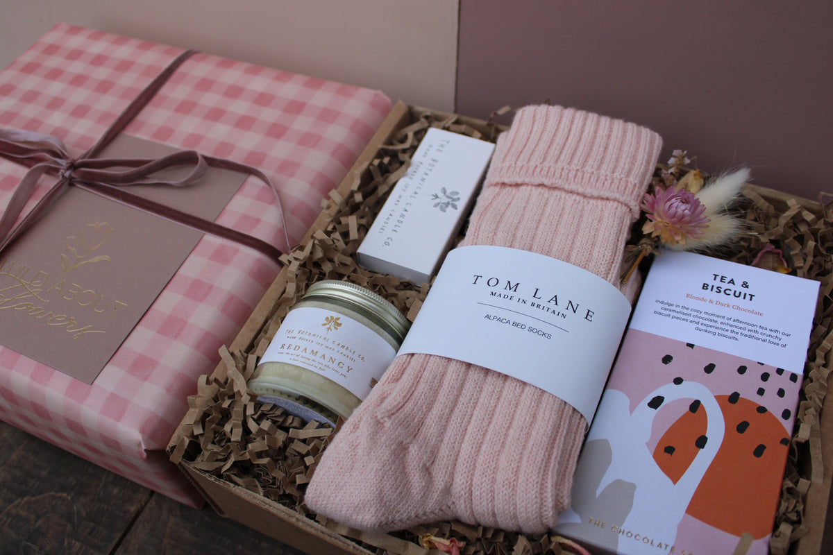 The 'Valentines Night in' Gift Box by Wild About Flowers includes alpaca wool bed socks, a candle, natural soap, dried flowers, and “Tea & Biscuit” chocolate—an indulgent romantic set presented on shredded brown paper.