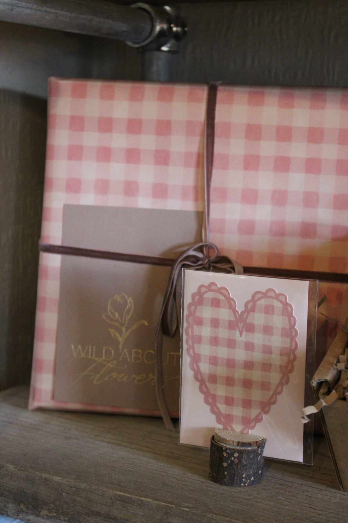 The 'Valentines Night in' Gift Box by Wild About Flowers, wrapped in pink and white checkered paper with a brown ribbon, sits beside a matching card with a checkered heart on a wooden shelf.