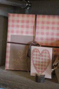 The 'Valentines Night in' Gift Box by Wild About Flowers, wrapped in pink and white checkered paper with a brown ribbon, sits beside a matching card with a checkered heart on a wooden shelf.
