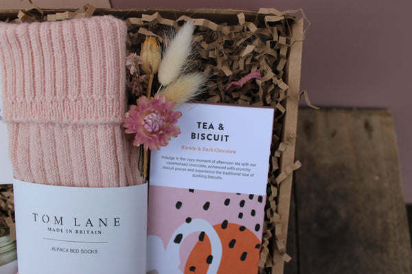 The Wild About Flowers 'Valentines Night in' Gift Box features pink alpaca wool bed socks, a "Tea & Biscuit" chocolate bar, dried flowers, and shred—all beautifully arranged for a romantic night in.