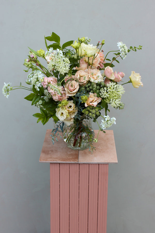 A stunning flower arrangement featuring pastel roses and lisianthus with lush green foliage is artfully presented in the Vintage Jug by Wild About Flowers, an eco-conscious gift elegantly resting on a pink pedestal against a plain light gray background.