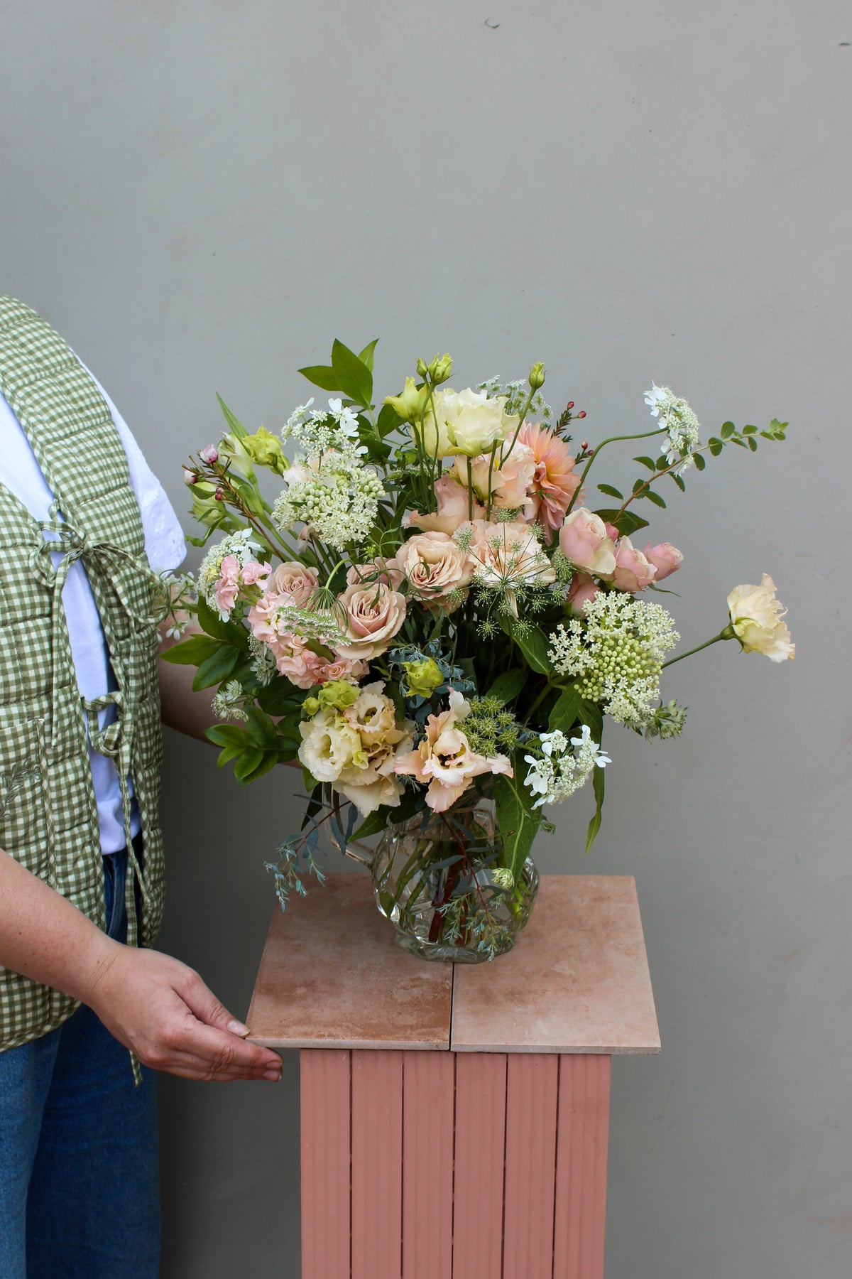 A person displays a captivating flower arrangement in the Vintage Jug by Wild About Flowers, featuring roses and lush greenery, presented on a pink pedestal. They wear a green checkered vest and denim pants against a light gray wall backdrop.