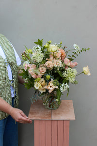 A person displays a captivating flower arrangement in the Vintage Jug by Wild About Flowers, featuring roses and lush greenery, presented on a pink pedestal. They wear a green checkered vest and denim pants against a light gray wall backdrop.