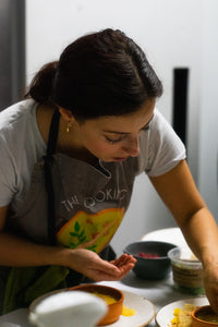 In "Supper Club with The Cooking Flea," a dark-haired woman in a gray apron and white shirt meticulously crafts plant-based cuisine. She sprinkles ingredients into a dish on the table, surrounded by bowls of fresh, seasonal produce.