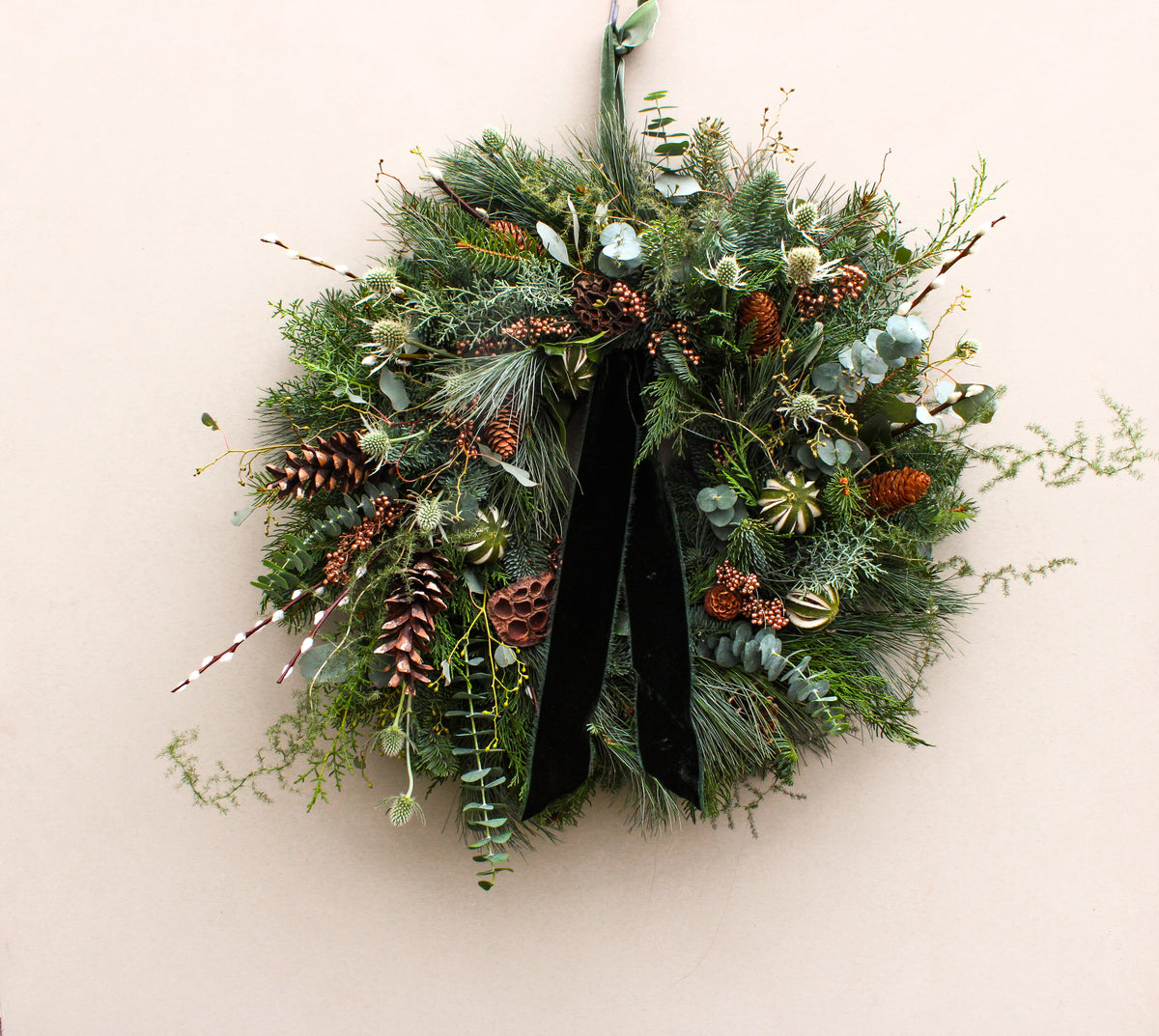Christmas wreath with greenery, pinecones, and a black ribbon on a beige wall.
