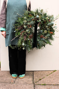 Person holding a festive wreath with greenery and pinecones against a plain background