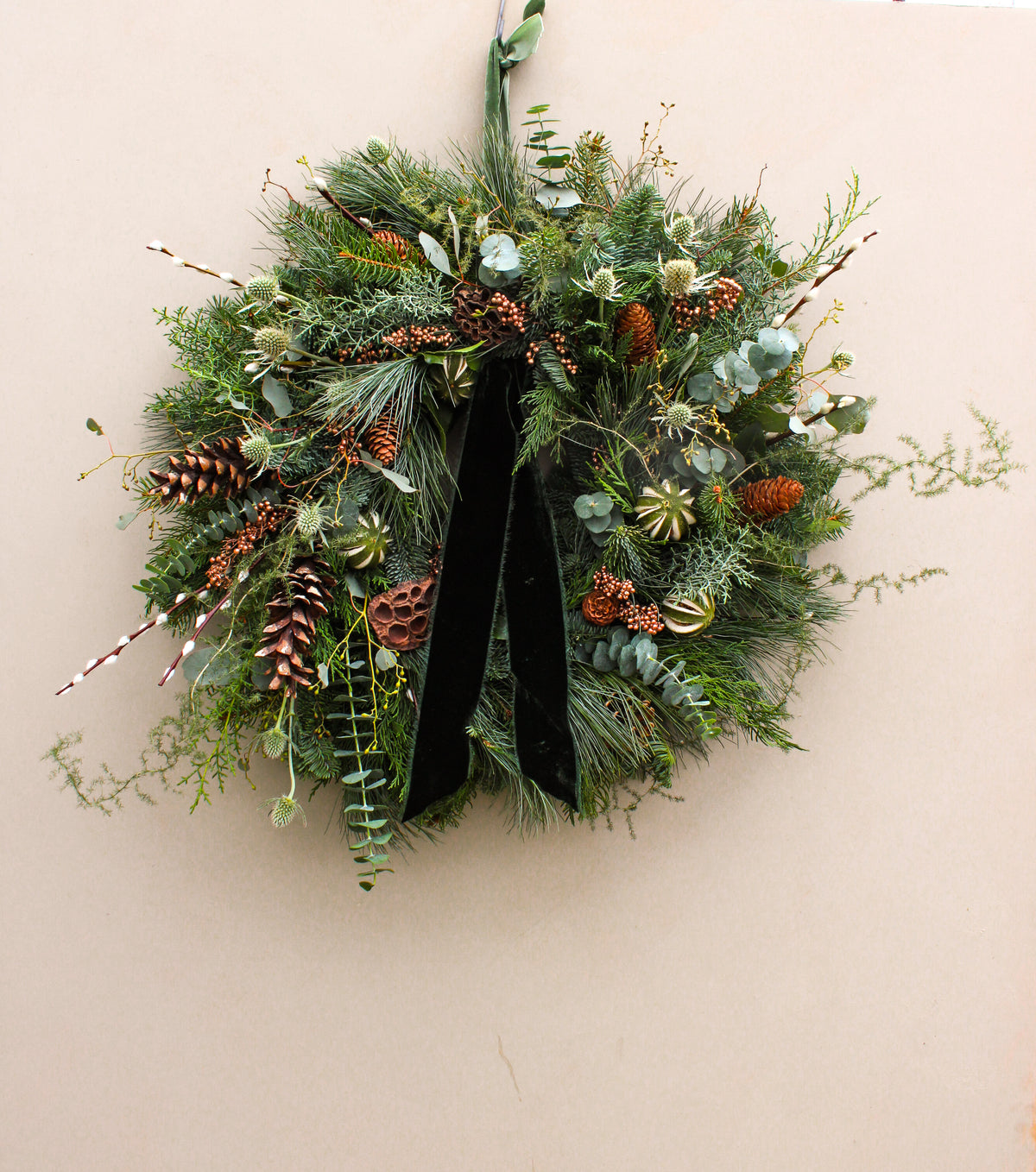 Decorative wreath with greenery, pinecones, and a black ribbon on a beige wall.