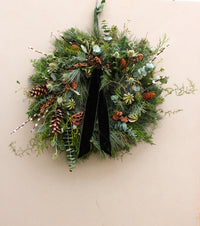 Decorative wreath with greenery, pinecones, and a black ribbon on a beige wall.