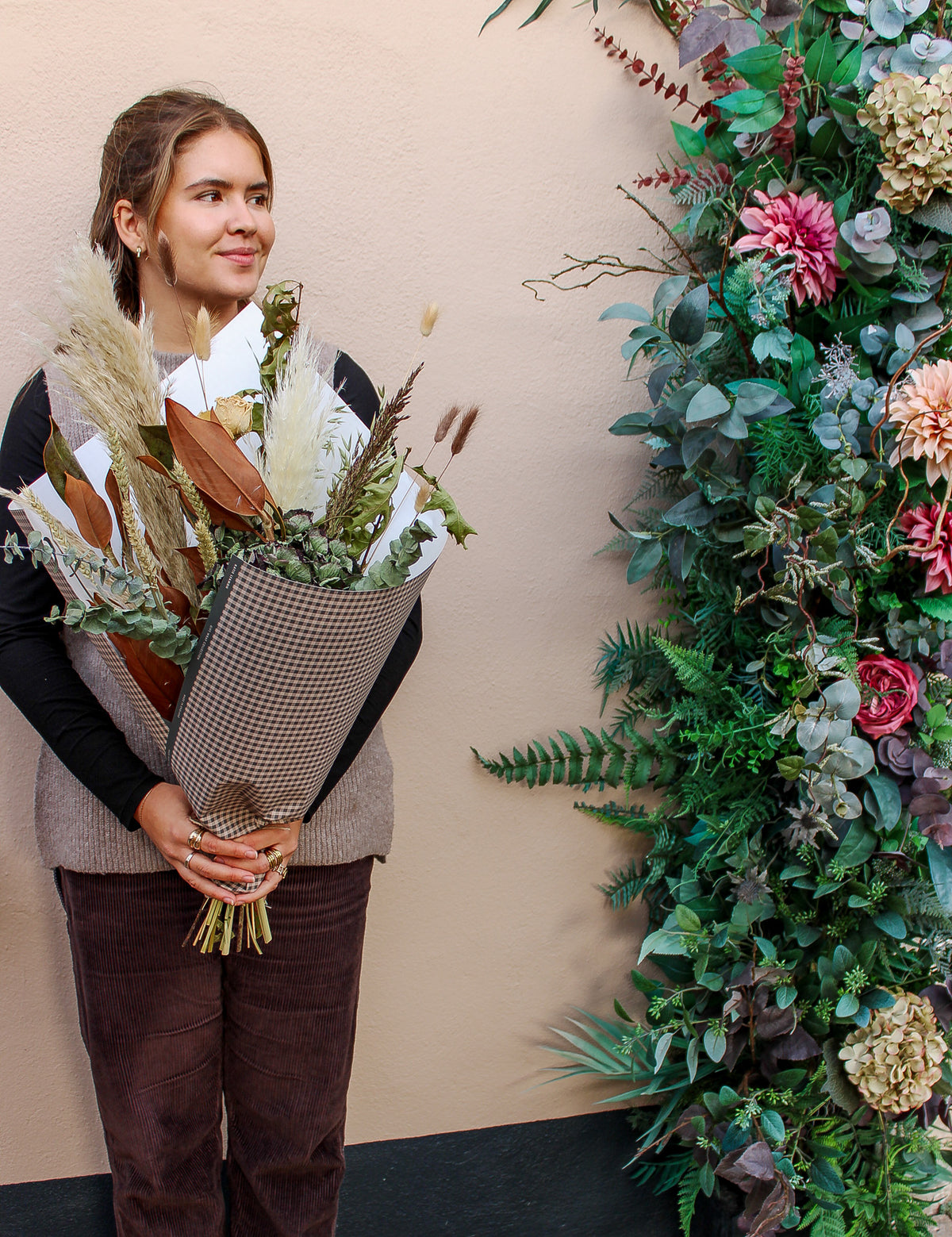 A woman smiles and looks to the side, holding the Wild About Flowers Pecan Pie bouquet, standing by a lush wall decorated with green leaves and pink flowers.