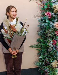 A woman smiles and looks to the side, holding the Wild About Flowers Pecan Pie bouquet, standing by a lush wall decorated with green leaves and pink flowers.