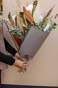 A person holds the Pecan Pie bouquet by Wild About Flowers, featuring dried and fresh pampas grass, eucalyptus, and brown leaves in black-and-white checkered paper—ideal for autumn flower lovers with local delivery available.