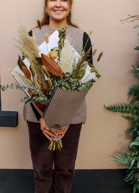 A woman holds the "Pecan Pie" bouquet by Wild About Flowers, wrapped in houndstooth-patterned paper, standing by a beige wall with green plants—highlighting the charm of local flower delivery.