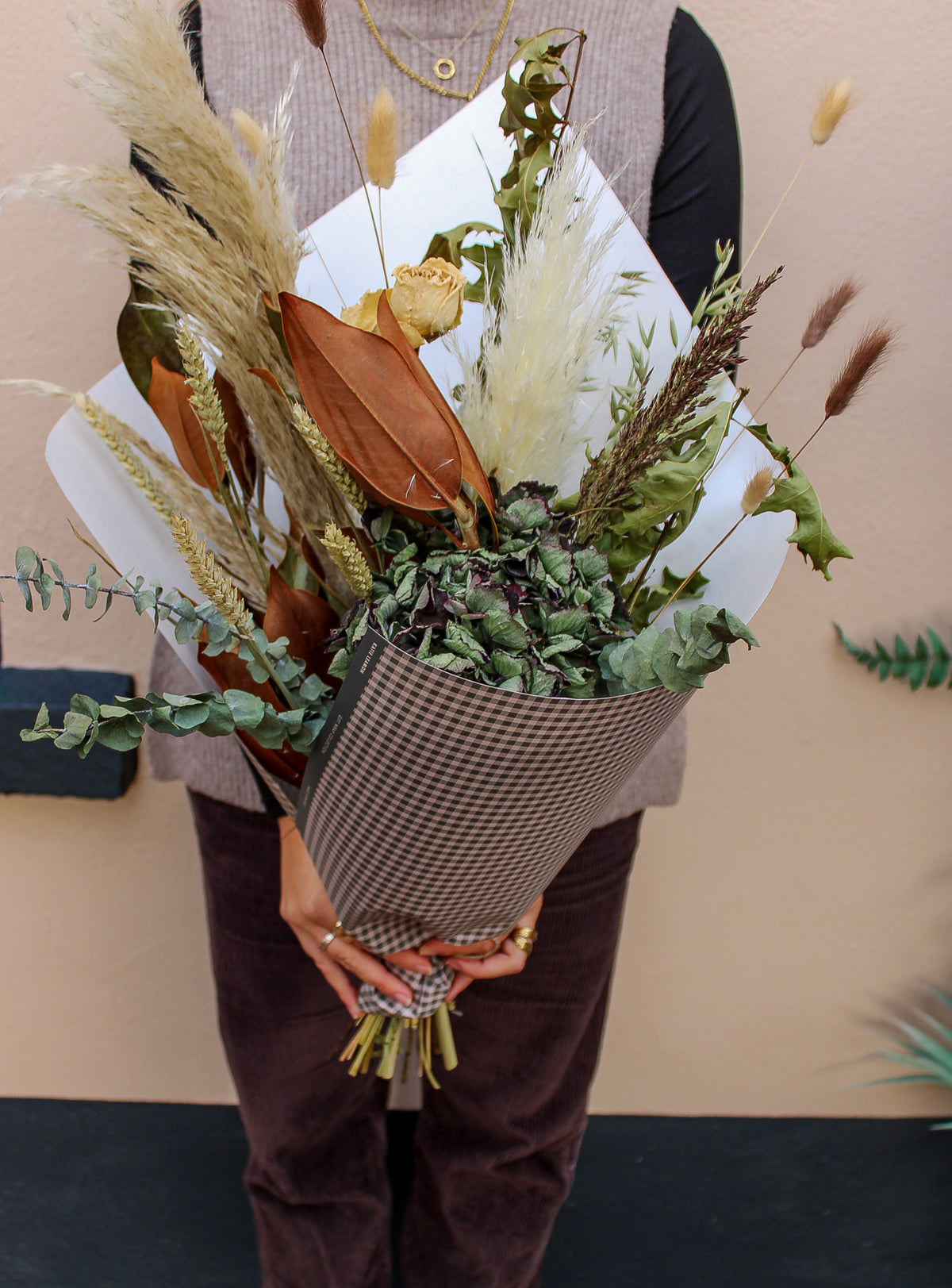 A person in a brown outfit holds the "Pecan Pie" dried flower bouquet by Wild About Flowers, featuring textured grasses, eucalyptus, and rich brown foliage, all wrapped in patterned paper against a light beige wall.