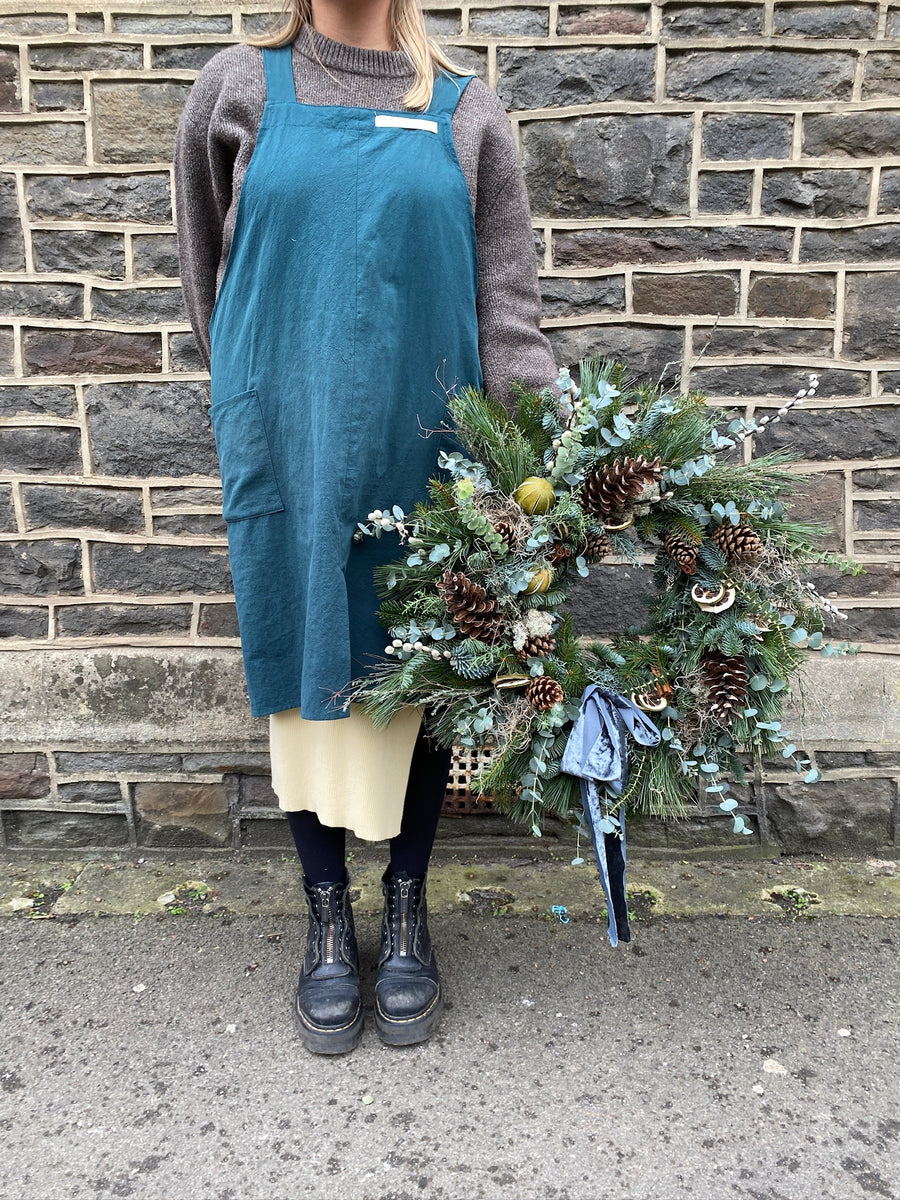 Person wearing a teal apron holding a large festive wreath with pine cones and greenery against a brick wall
