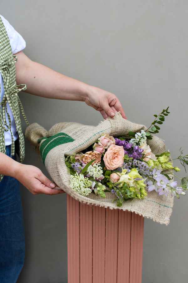 A person showcases a "Coffee wrap" bouquet by Wild About Flowers, featuring seasonal pink and peach roses, purple and white blooms, and lush green foliage set against a gray backdrop.