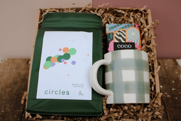 The 'Love in a mug' Gift Box by Wix includes a sustainable collection featuring a green plaid ceramic mug, a bag of Circles Brazilian Espresso coffee with a white label adorned with colorful dots, and a small, vibrant package marked "COCO." All items are carefully cushioned in shredded brown paper.