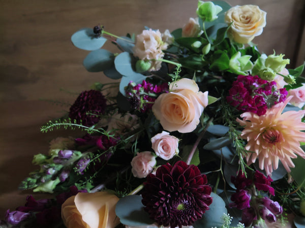 The 'From the Heart' Tied Sheaf by Wild About Flowers features a vibrant bouquet composed of peach roses, dark red chrysanthemums, pink carnations, and green foliage set against a wooden background. This eco-friendly arrangement is stylishly lush and cascading, showcasing a commitment to sustainable design.