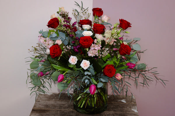 The Valentine's Day Blooms bouquet by Wild About Flowers is a romantic gift, featuring red roses, pink and white blooms, and purple tulips in a glass vase. Lush greenery complements the arrangement on a rustic wooden surface against a two-toned wall—a perfect display for Valentine's Day.