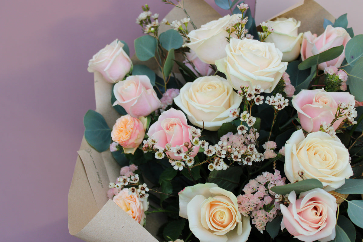 The 'All About the Rose' Bouquet by Wix is a perfect Mother's Day gift, featuring white and pale pink roses surrounded by sprigs of small white flowers and green leaves, all elegantly wrapped in brown paper. The background of the arrangement is a soft, dusty pink color. Ideal for local delivery in Bristol.