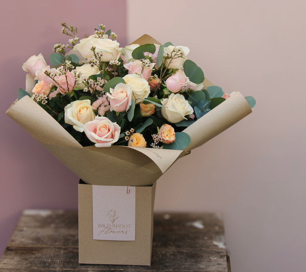 For Mother's Day, the 'All About the Rose' Bouquet by Wix features roses in delicate shades of white, peach, and pale pink. The arrangement is elegantly wrapped in brown paper and placed in a brown box labeled "Wild About Flowers." It sits beautifully on a wooden table with a pink and beige background, making it perfect for local delivery in Bristol.