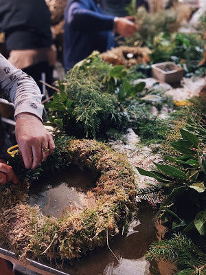 An individual is assembling a wreath using an assortment of green leaves and plant materials at a table. In the background, others are also participating in the Wreath Workshop by Event, crafting their own festive wreaths. The table is adorned with sustainable foliage and various crafting tools.