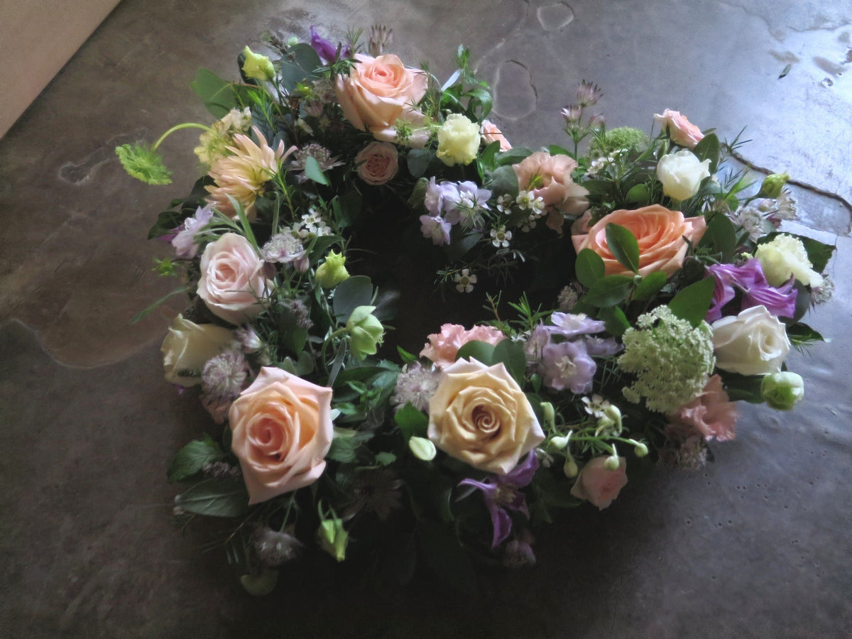 The 'From the Garden' Heart Design by Wix lies on a dark surface, serving as a tribute to your loved one. This heart-shaped floral arrangement features a seasonal mix of roses, including pink and yellow varieties, complemented by white and purple blooms and green foliage. The colors and arrangement create a vibrant and elegant display.