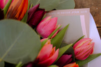 The Armful of Tulip Bouquet by Wix includes vibrant pink, orange, and dark purple tulips nestled among lush greenery, complete with a partially visible card. Perfect for Valentine's Day, this romantic gift elegantly rests on a wooden surface.