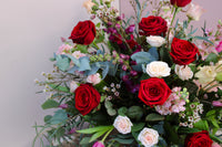 The Valentine's Day Blooms by Wild About Flowers features a vibrant bouquet of red and white roses, pink flowers, and green foliage arranged in a vase against a neutral background—an elegant display perfect for a romantic gift.