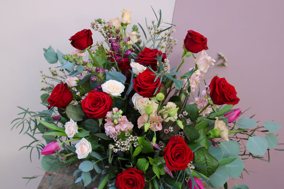 The Valentine's Day Blooms by Wild About Flowers is a vibrant vase bouquet with red roses, white roses, pink tulips, and assorted greenery set against a pale pink and light grey background—ideal for a romantic Valentine’s Day gift.