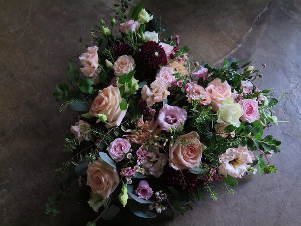 The 'From the Heart' Heart Design by Wild About Flowers is a heart-shaped floral tribute featuring pink roses, light pink and white flowers, and dark maroon accents. It is lush with greenery and scented herbs, all set against a dark, textured background.