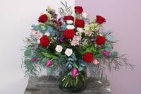 The "Valentine's Day Blooms" by Wild About Flowers graces the wooden table with a vibrant bouquet of red roses, pink lisianthus, small white roses, greenery with eucalyptus sprigs, and purple tulips—a perfect romantic gift against a two-toned pink and mauve wall.