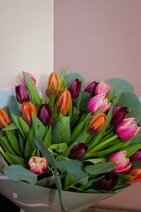 The Armful of Tulip Bouquet by Wix showcases vibrant tulips in pink, orange, and deep purple hues. Accented with lush green leaves against a pink and beige backdrop, it's an ideal romantic gift.