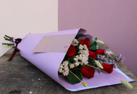 The "Early (love) bird" bouquet by Wix, featuring red roses and elegant white flowers wrapped in purple paper with a decorative card, rests gracefully on a wooden surface against light and dark pink paneling.