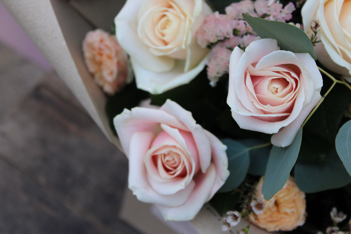 A close-up image of the 'All About the Rose' Bouquet by Wix shows a stunning Mother's Day arrangement featuring pale pink and white roses. Surrounding the roses are sprigs of green leaves and small pale pink flowers, all arranged in a soft, elegant composition. Perfect for local delivery in Bristol, the bouquet is set against a neutral background.