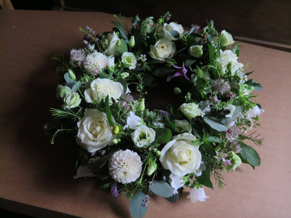 A lush and elegant floral wreath composed of white roses, cream-colored dahlias, and various greenery is meticulously arranged on a brown surface. The flowers and foliage are intricately woven into a circular shape. Set against a dimly lit background, this timeless tribute by Wix is beautifully showcased under the product name Wild About Wreath.