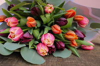 The Armful of Tulip Bouquet by Wix features vibrant pink, orange, and deep purple blooms with lush green leaves. Elegantly arranged on a wooden surface, this romantic gift perfectly captures the essence of love and beauty.