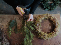 A person seen from above is crafting a sustainable foliage wreath on a wooden table during an Event Wreath Workshop. They are cutting evergreen branches with yellow-handled scissors. On the table are branches, a partially completed moss wreath, and some loose twigs and greenery, capturing the essence of this festive workshop.