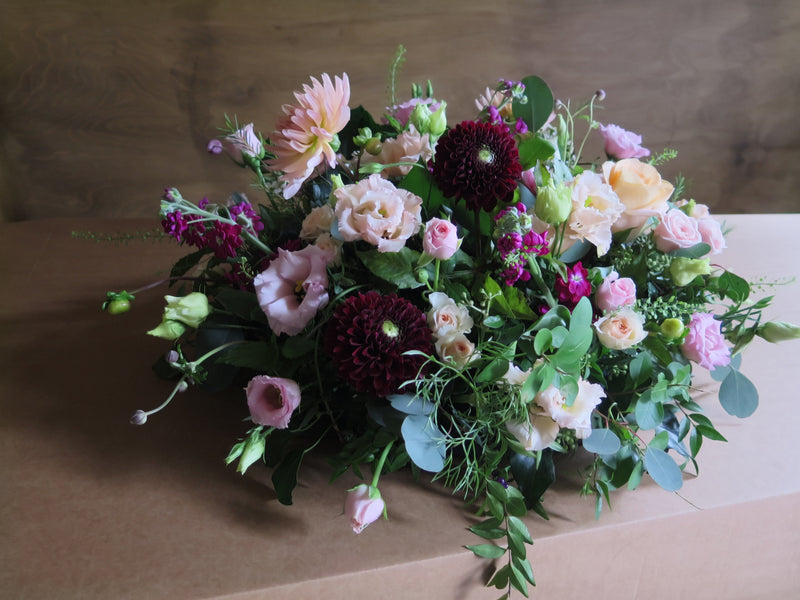 The 'From the Heart' Posy Design by Wix features a lush floral arrangement that includes pink roses, dark red dahlias, light pink lisianthus, and greenery. Set on a cardboard surface against a wooden backdrop, the varying textures and colors create a vibrant, natural tribute perfect for a final farewell.