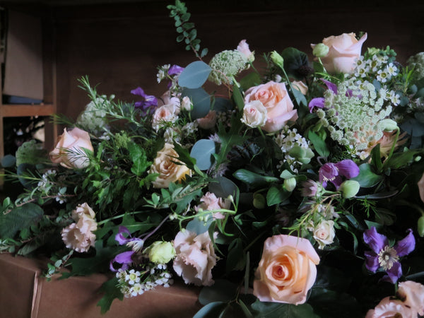 The 'From the Garden' Casket Spray by Wild About Flowers is an elegant arrangement of seasonal blooms, featuring pale pink roses, green foliage, and purple accents beautifully set against a dark background.