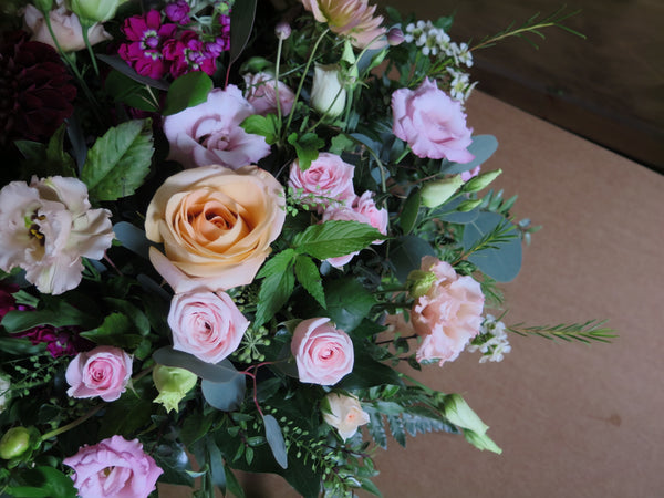 The 'From the Heart' Posy Design by Wix features a vibrant arrangement of various flowers, including pale orange roses, light pink roses, purple blooms, and white buds, all surrounded by fresh cut foliage and lush green leaves. The floral display is elegantly set against a brown cardboard background.