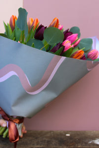 The Armful of Tulip Bouquet by Wix features vibrant tulips in pink, orange, and purple shades. Wrapped in wavy decorative paper and tied with a ribbon against a soft pink background, it's an ideal romantic gift for any occasion.