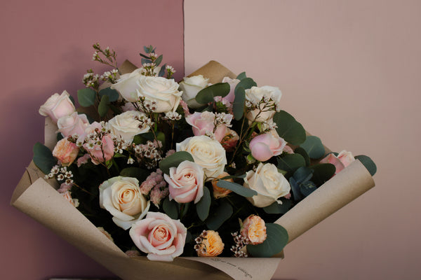 Introducing the 'All About the Rose' Bouquet by Wix: A Mother's Day arrangement featuring white and pale pink roses, complemented by small white blossoms and green leaves, all wrapped in elegant brown paper. The bouquet is set against a backdrop of muted pink and beige tones. Available for local delivery in Bristol.