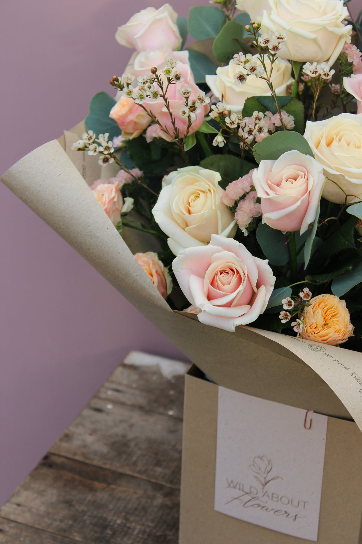 The 'All About the Rose' Bouquet by Wix features a stunning arrangement of cream and pale pink roses, accented with delicate small white filler flowers and charming peach-colored buds, all elegantly wrapped in brown paper. Perfect for Mother's Day, this bouquet arrives in a box labeled "Wild about flowers" and is beautifully displayed on a rustic wooden surface. Local delivery is available in Bristol.