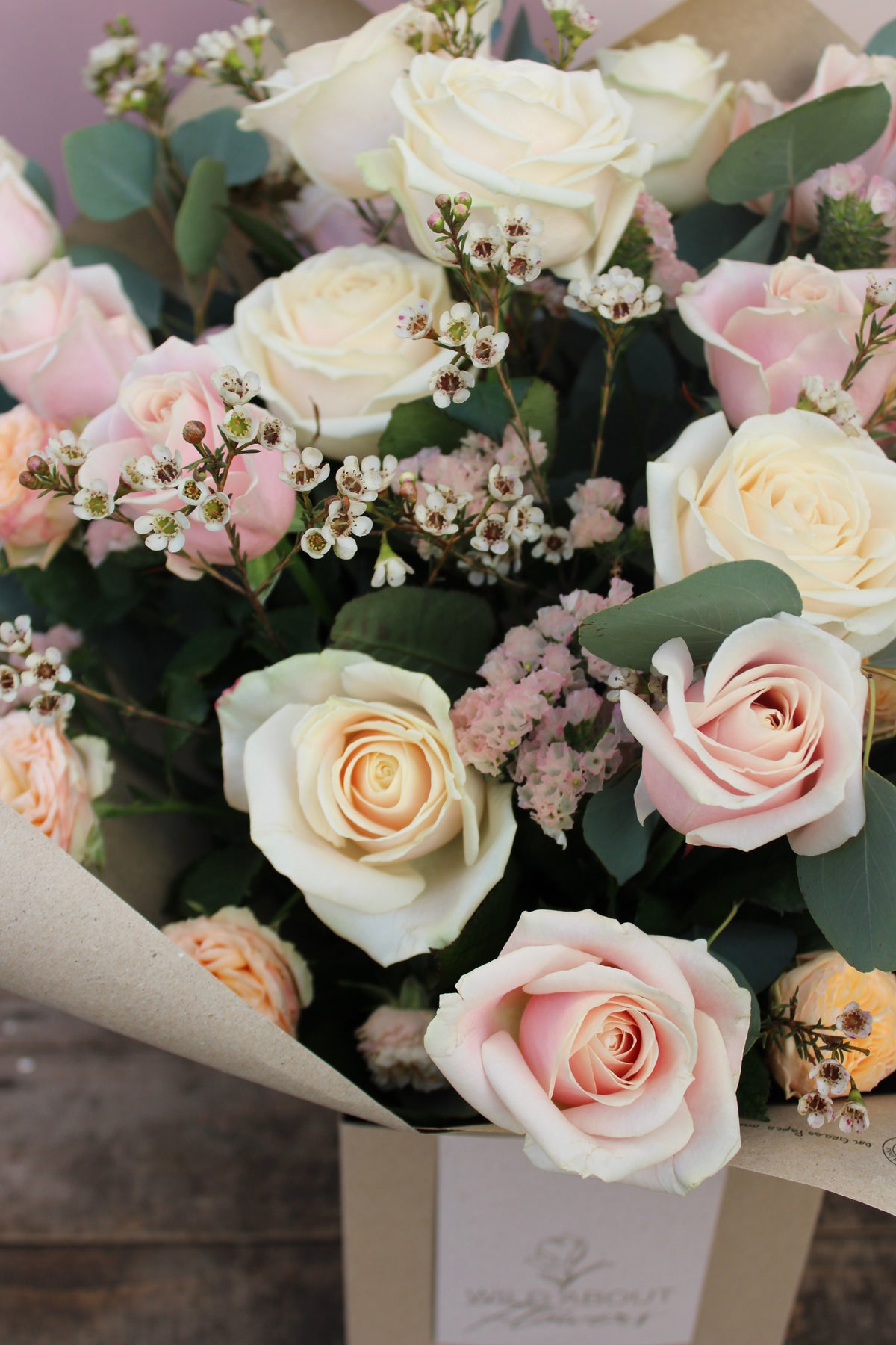 Introducing the 'All About the Rose' Bouquet by Wix, a stunning Mother's Day arrangement featuring a blend of white, cream, and light pink roses intermingled with delicate waxflowers and lush green foliage. This elegant bouquet is wrapped in natural-colored paper and displayed on a wooden surface. Now available for local delivery in Bristol.