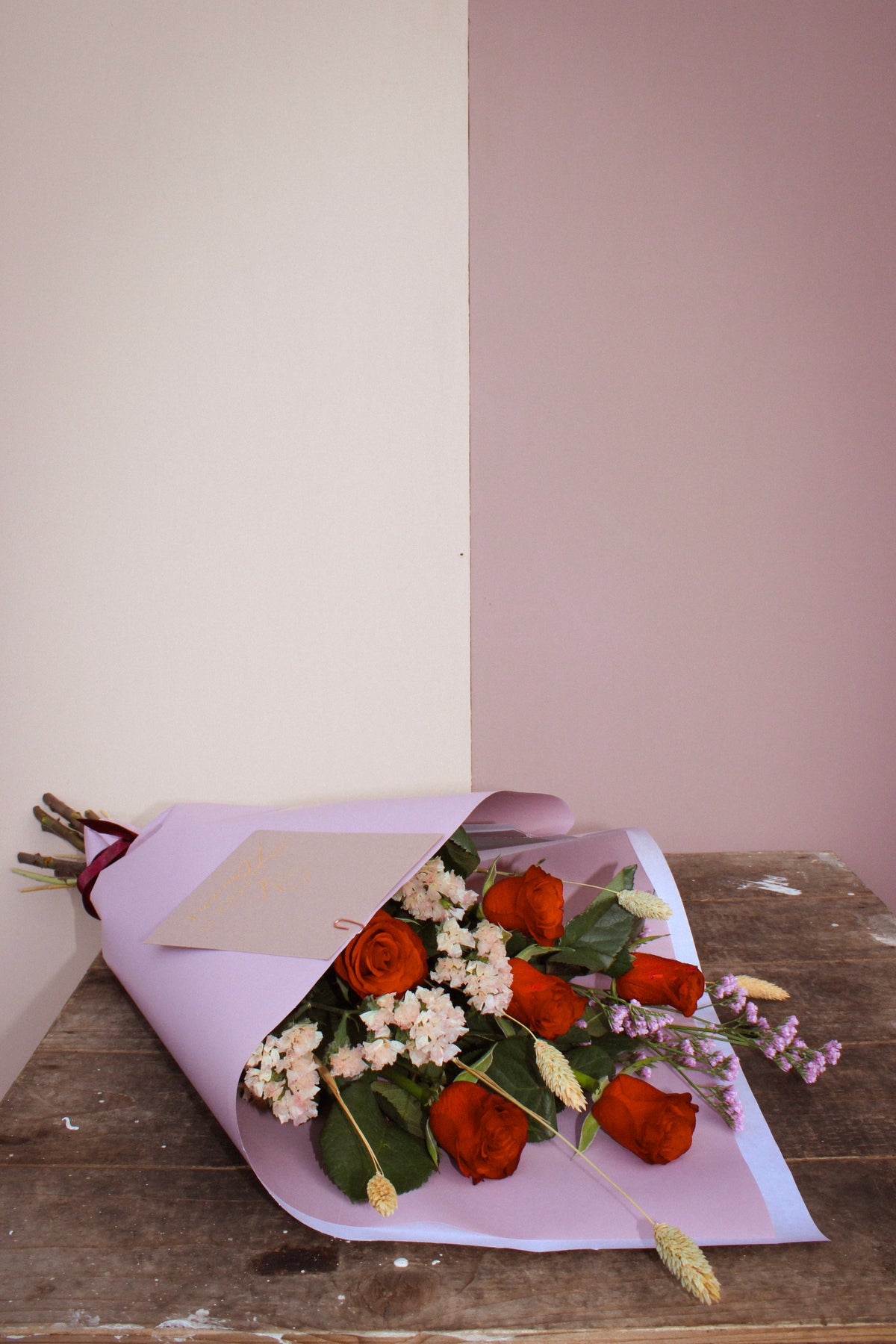 The Wix "Early (love) bird" bouquet, featuring orange and red roses wrapped in lavender paper with pink and white flowers, rests on a rustic wooden surface. The floral arrangement beautifully contrasts against an off-white and mauve wall.