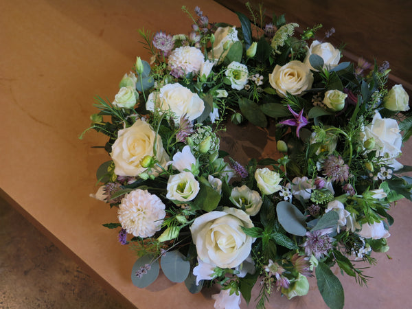 The Wild About Wreath by Wild About Flowers is an elegant arrangement featuring white roses, dahlias, and lush greenery on a wooden surface. Interwoven with various leaves and smaller flowers, it serves as a beautiful tribute for remembrance.